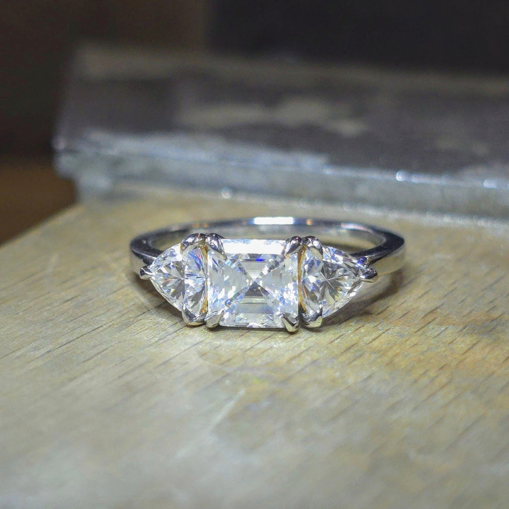 Trendig platinaring med tre gnistrande moissaniter – cushion‑cut mittsten, två trillionslipade sidostenar, och infattning i rödguld