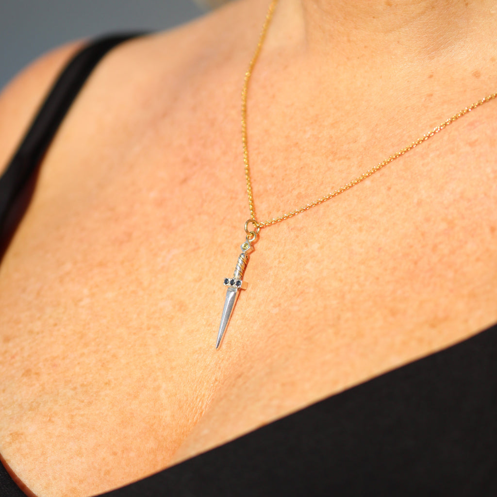 Ett hängsmycke formad som en liten silversvärd med en guldkedja. Svärdets fäste är prytt med svarta ädelstenar, och en ljus sten sitter högst upp på handtaget.