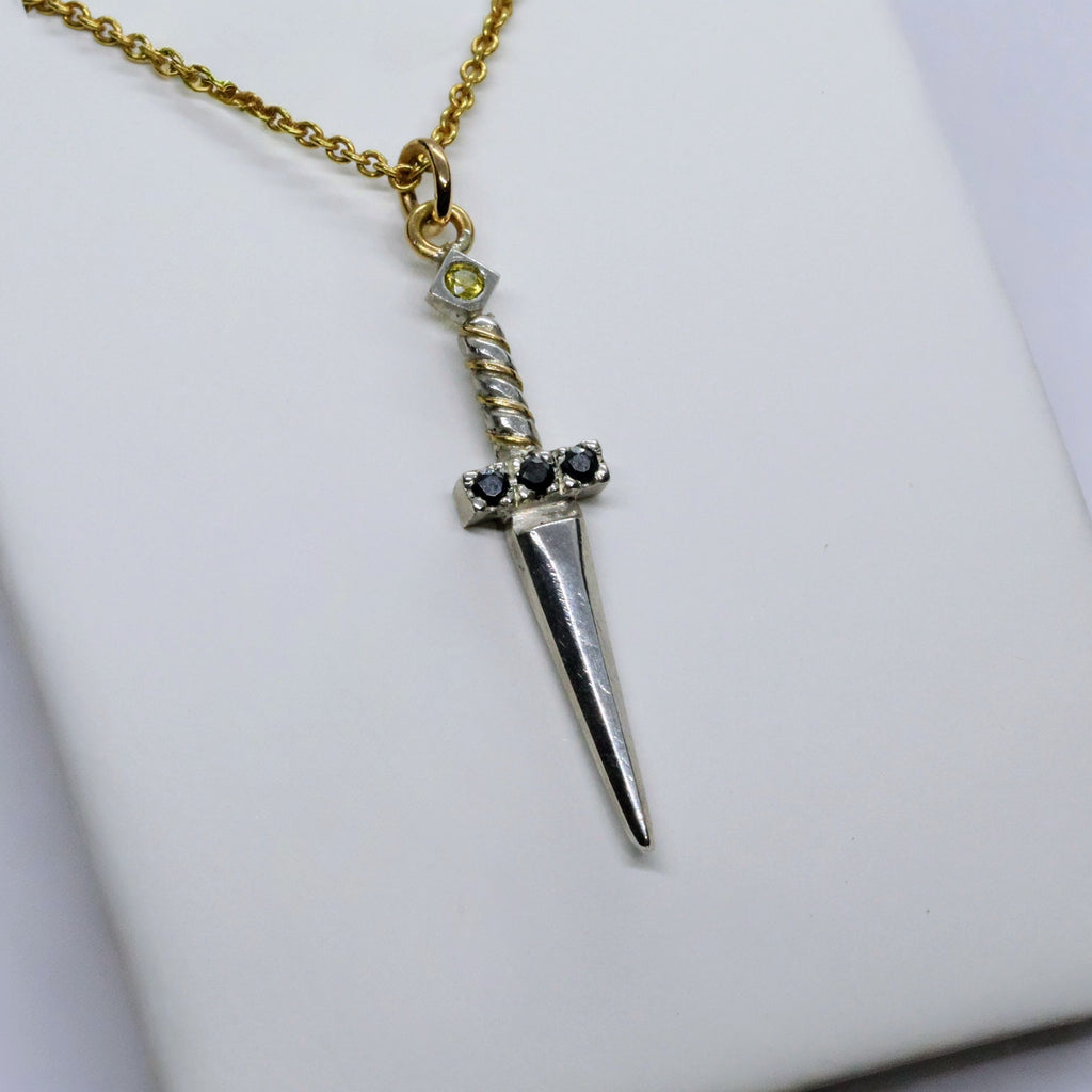 Ett hängsmycke formad som en liten silversvärd med en guldkedja. Svärdets fäste är prytt med svarta ädelstenar, och en ljus sten sitter högst upp på handtaget.