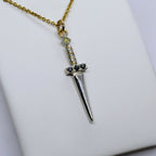 Ett hängsmycke formad som en liten silversvärd med en guldkedja. Svärdets fäste är prytt med svarta ädelstenar, och en ljus sten sitter högst upp på handtaget.