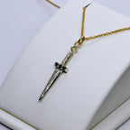 Ett hängsmycke formad som en liten silversvärd med en guldkedja. Svärdets fäste är prytt med svarta ädelstenar, och en ljus sten sitter högst upp på handtaget.