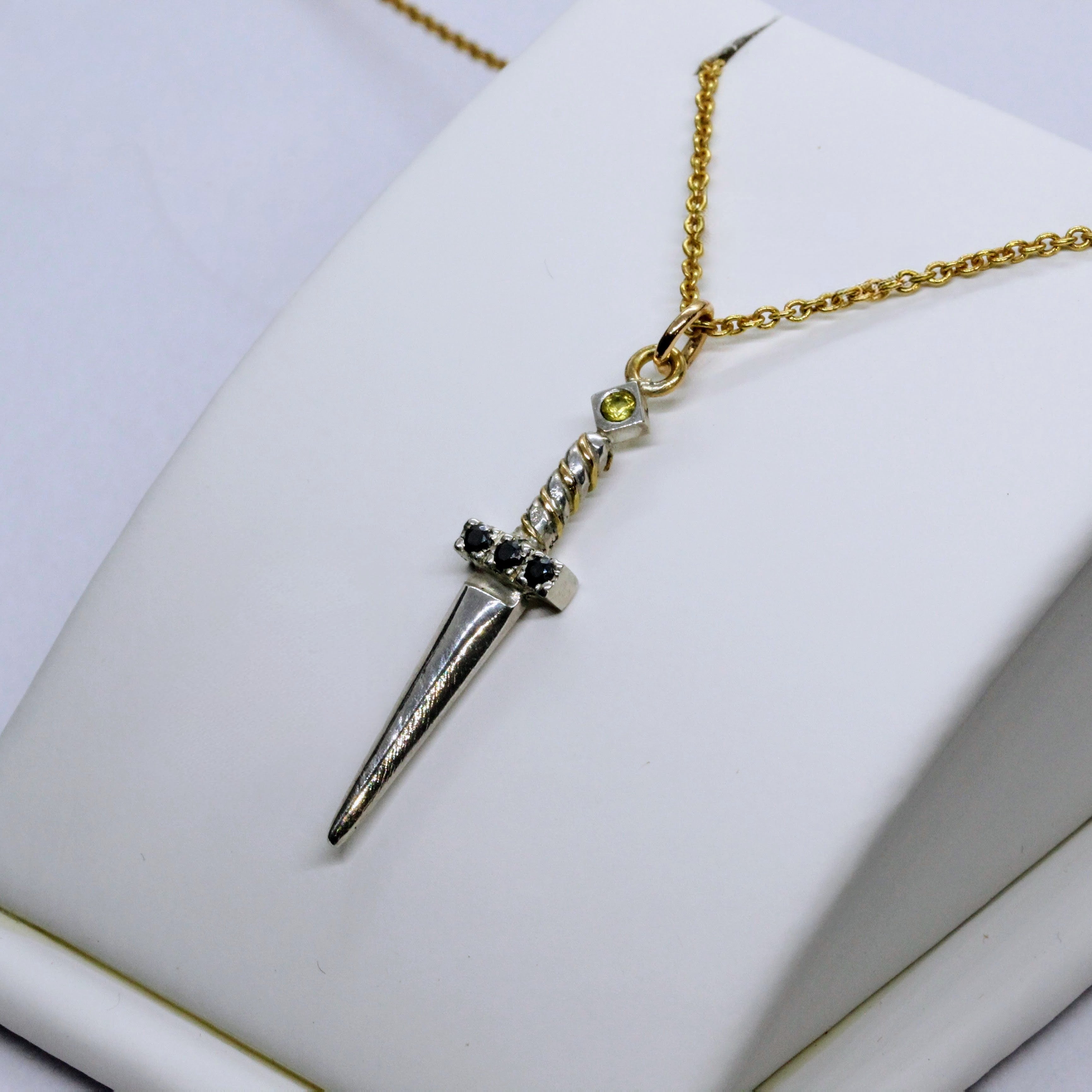 Ett hängsmycke formad som en liten silversvärd med en guldkedja. Svärdets fäste är prytt med svarta ädelstenar, och en ljus sten sitter högst upp på handtaget.